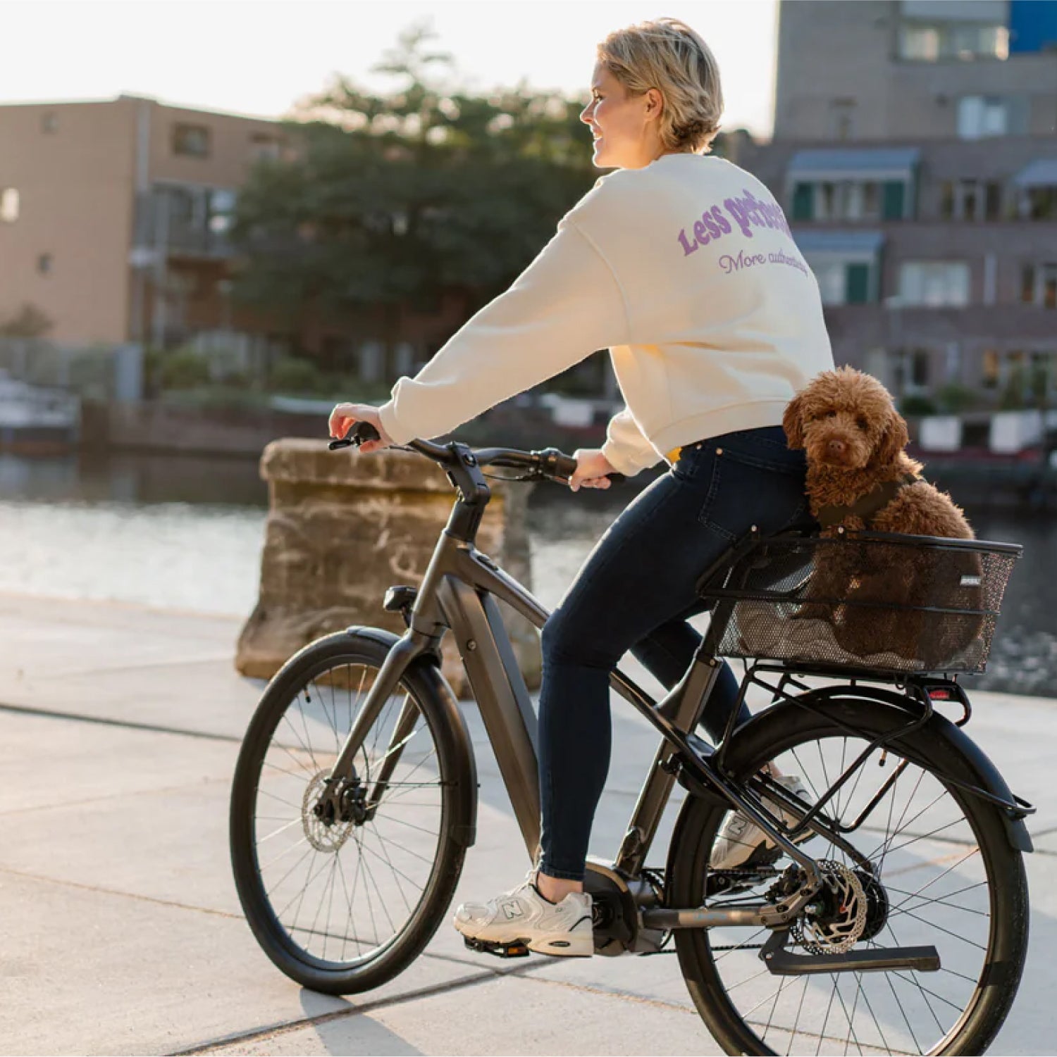 Blonde lachende vrouw op donkergrijze Levante E4 e-bike voor water met bruin hondje achterop in metalen fietsmandje op bagagedrager